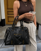 Black - A person wearing a brown ribbed tank top and cream wide-leg pants carries a large black leather handbag with gold-tone hardware and fringe details while holding an iced coffee outdoors.