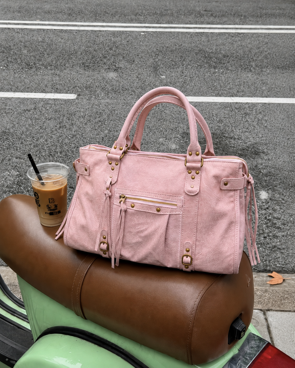 Pink - A light pink canvas tote bag with gold-tone hardware and leather straps, resting on a brown leather seat of a mint green scooter next to an iced coffee on an asphalt street.