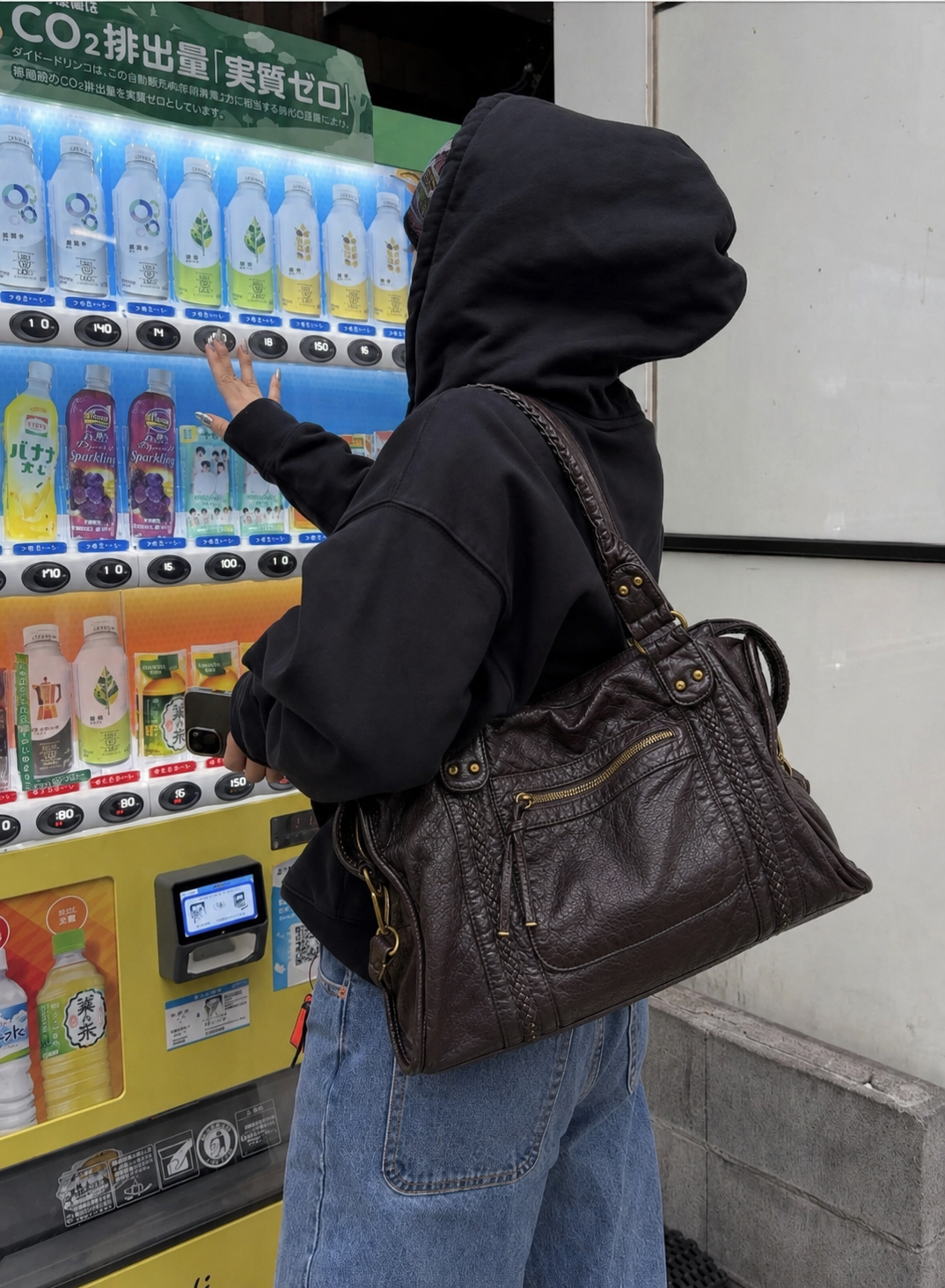 Coffee - A person in a black hoodie and blue jeans stands in front of a vending machine, carrying a large dark brown leather shoulder bag with gold-tone hardware.