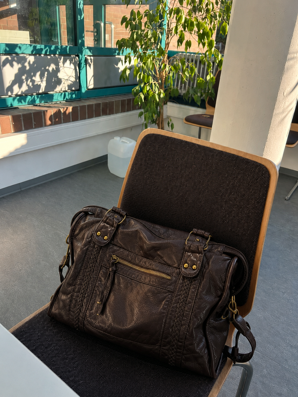 Coffee - A dark brown leather duffel bag with gold-tone hardware and multiple zippered pockets, resting on a dark upholstered office chair in a modern indoor setting.