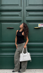 White - A woman posing in front of a dark green wooden door, wearing a black sleeveless top, wide-leg gray cargo pants, and holding a white handbag.